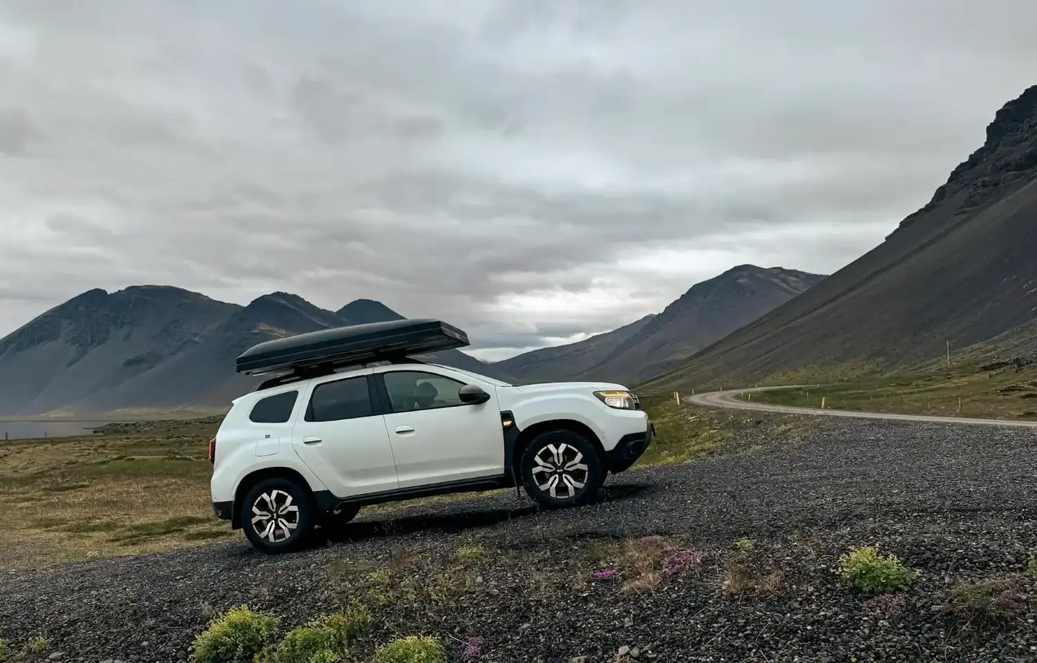 White SUV with a rooftop cargo box parked beside a winding road in rural Iceland, surrounded by dark volcanic mountains under an overcast sky.