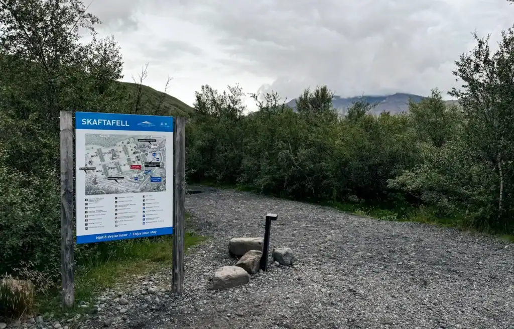 Trailhead with a map sign for Skaftafell in Iceland, surrounded by bushes and a gravel path under cloudy skies.