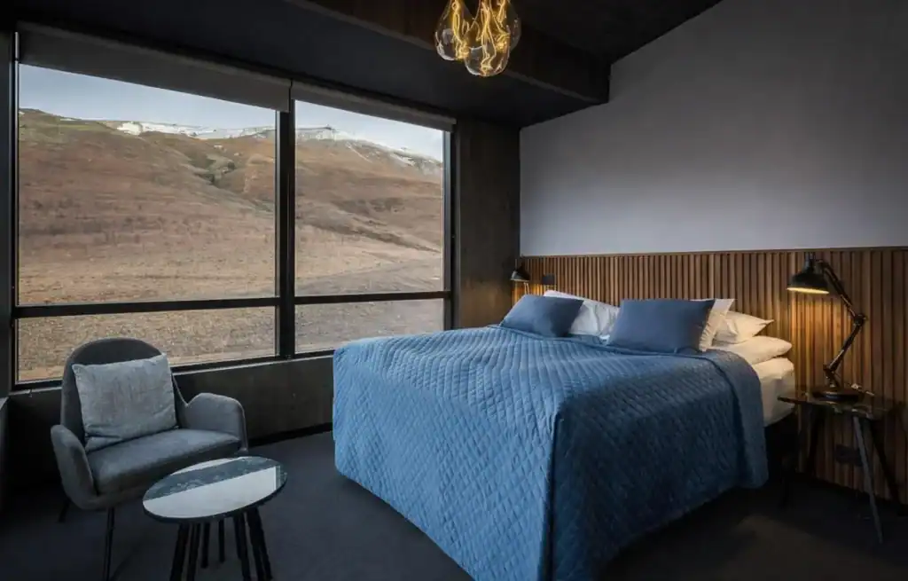 Modern bedroom with large window overlooking a mountainous landscape, blue bedspread, and minimalist furniture.