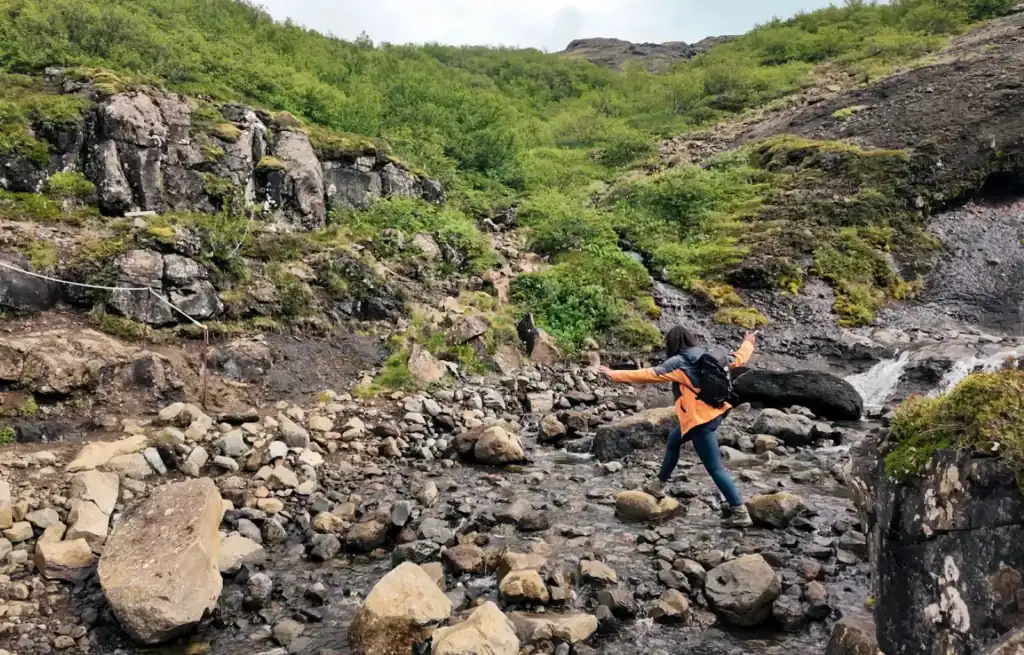 Hiker crossing a rocky stream with lush green hills and a small waterfall in the background.