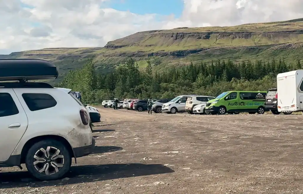 Cars parked on a gravel lot with green hills and cliffs in the background under a partly cloudy sky.