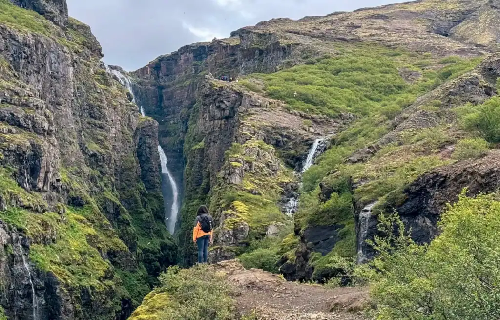 Hiker standing on a rocky trail overlooking a deep canyon with waterfalls and lush green vegetation.
