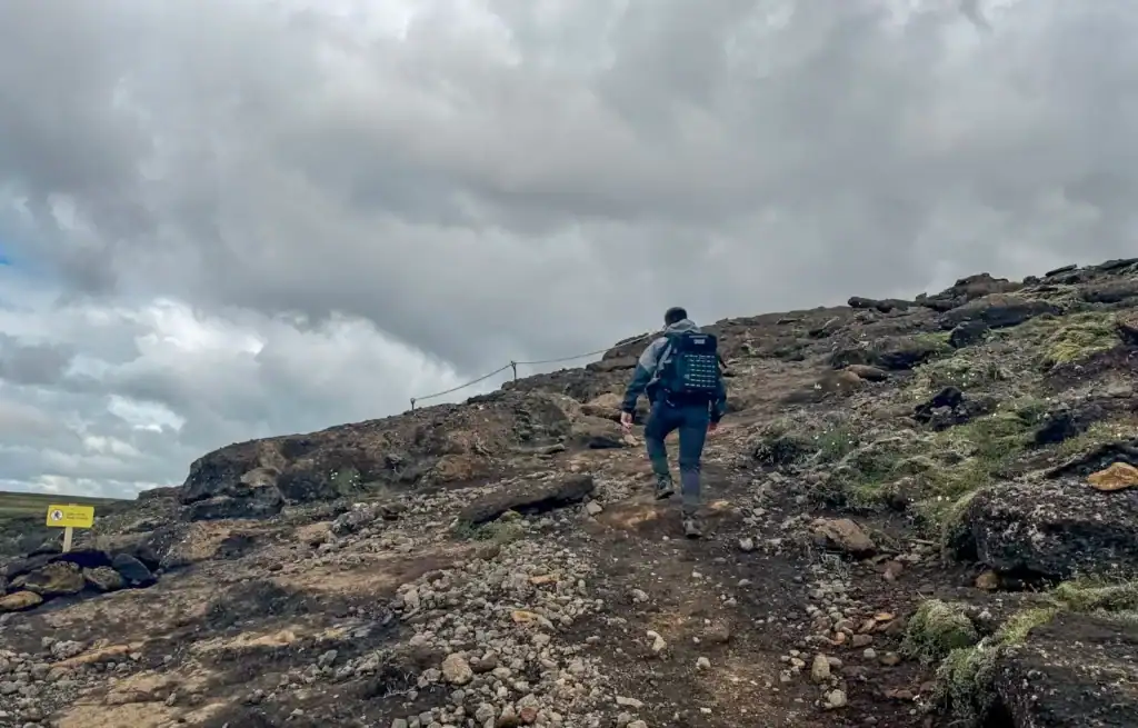 Hiker ascending a rocky trail under cloudy sky in a rugged landscape.