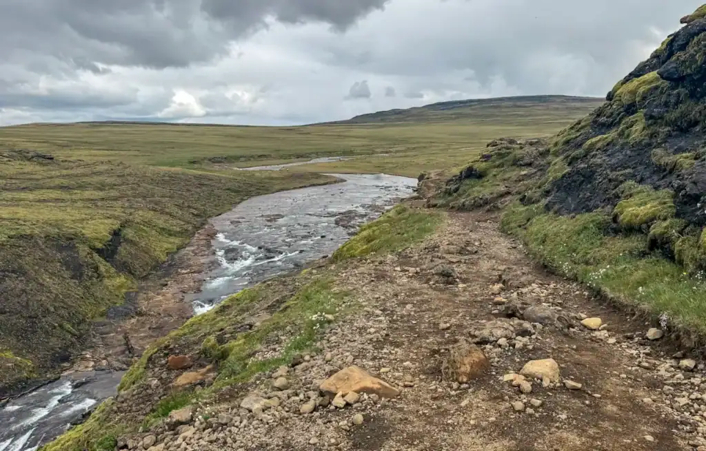 Rugged dirt trail along a river in a green Icelandic landscape under cloudy sky.