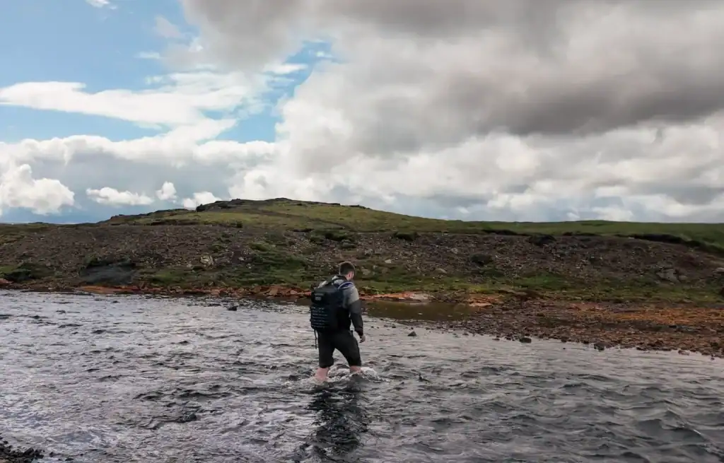 Hiker wading through a river with a backpack, green hills and cloudy sky in the background.