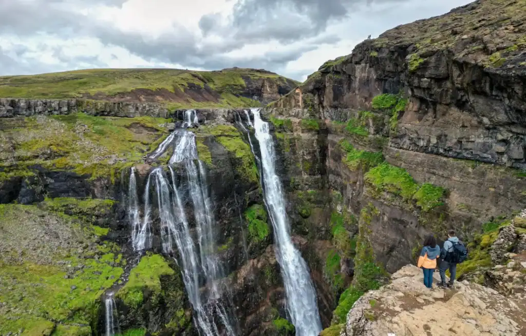 Two hikers stand on a rocky ledge overlooking the cascading Glymur Waterfall in Iceland.