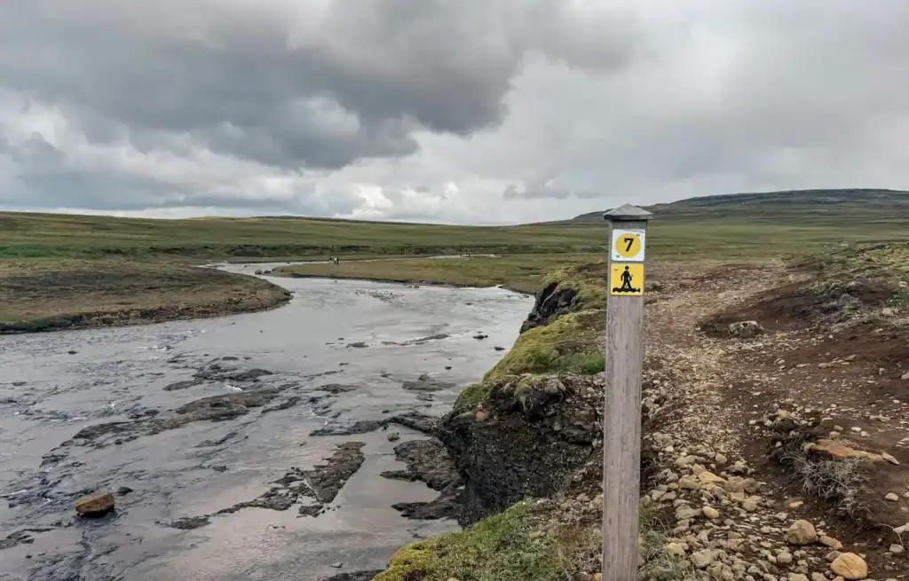 River flowing through a grassy landscape with a trail marker post and cloudy sky.