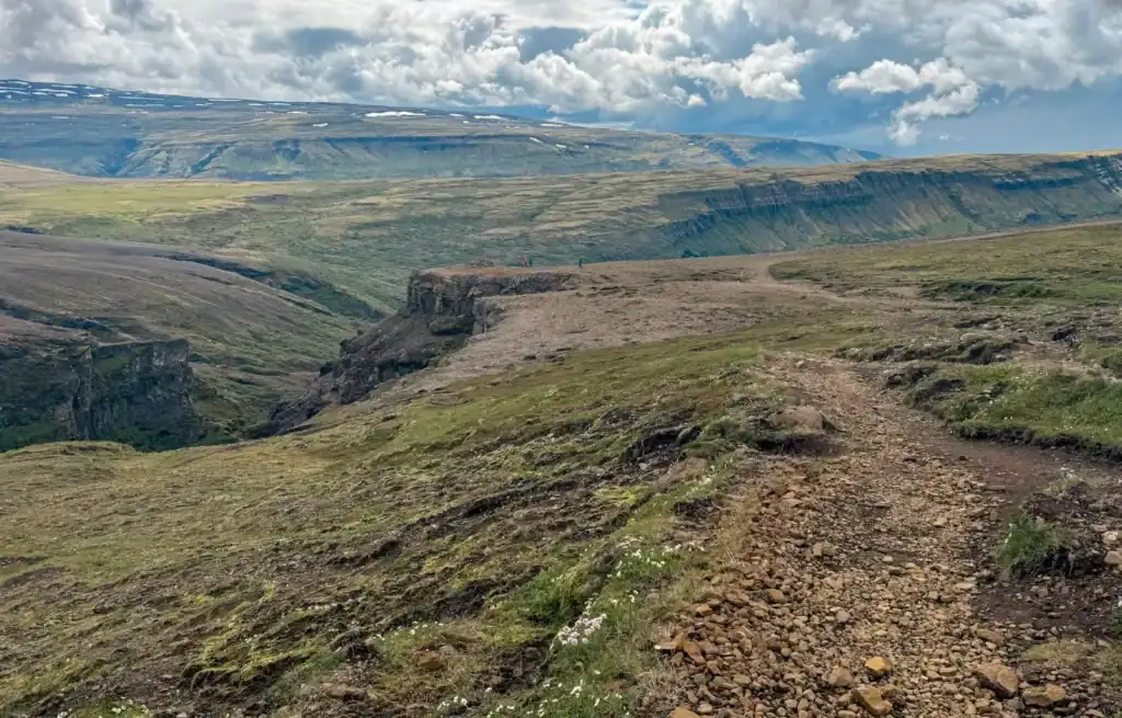 Rugged trail along a grassy cliff edge overlooking a deep canyon and distant mountains under cloudy skies.
