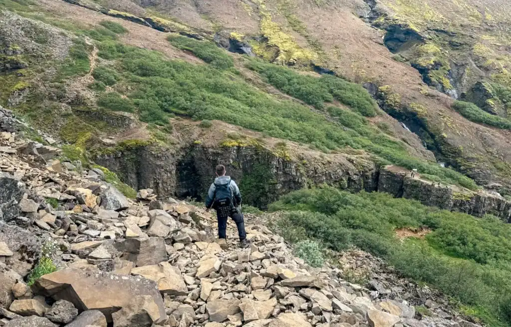 Hiker with backpack walking on rocky trail in lush green landscape with steep hillside.