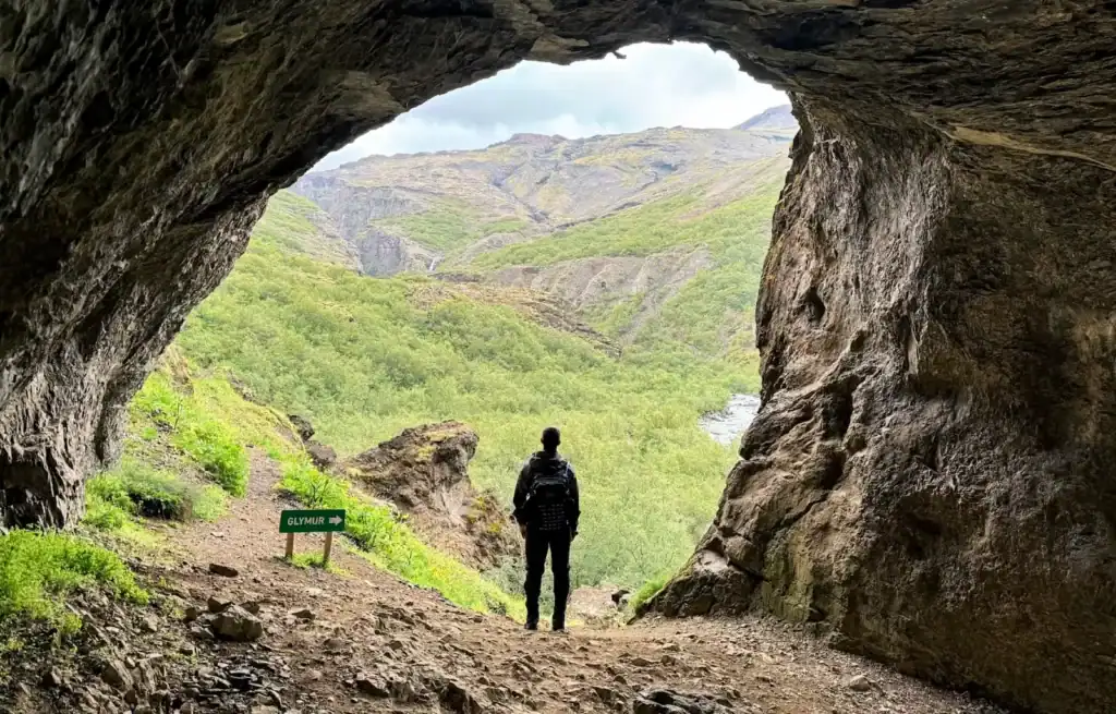 Hiker standing at the entrance of a cave with lush green mountains in the background.