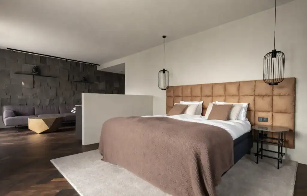 Modern hotel bedroom with a large bed, beige tufted headboard, and pendant lights, with a sitting area in the background.