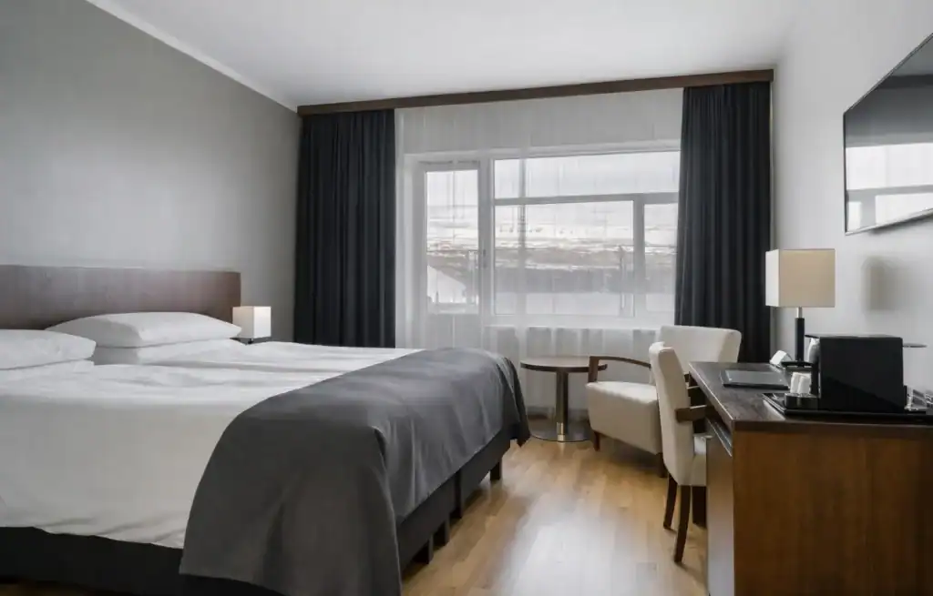 Modern hotel room with a large bed, window with dark curtains, and a desk with a chair and lamp.