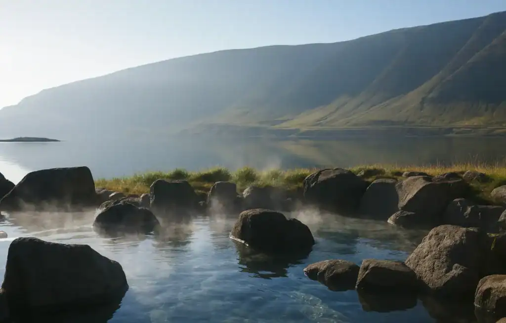 Steam rises from hot springs with large rocks, overlooking a calm lake and green mountains in the distance.