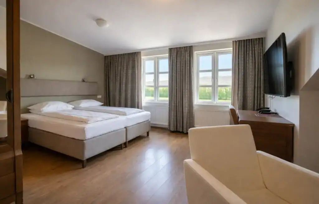 Spacious hotel room with a large bed, two windows with curtains, a TV on the wall, and a sitting area with a cream armchair.