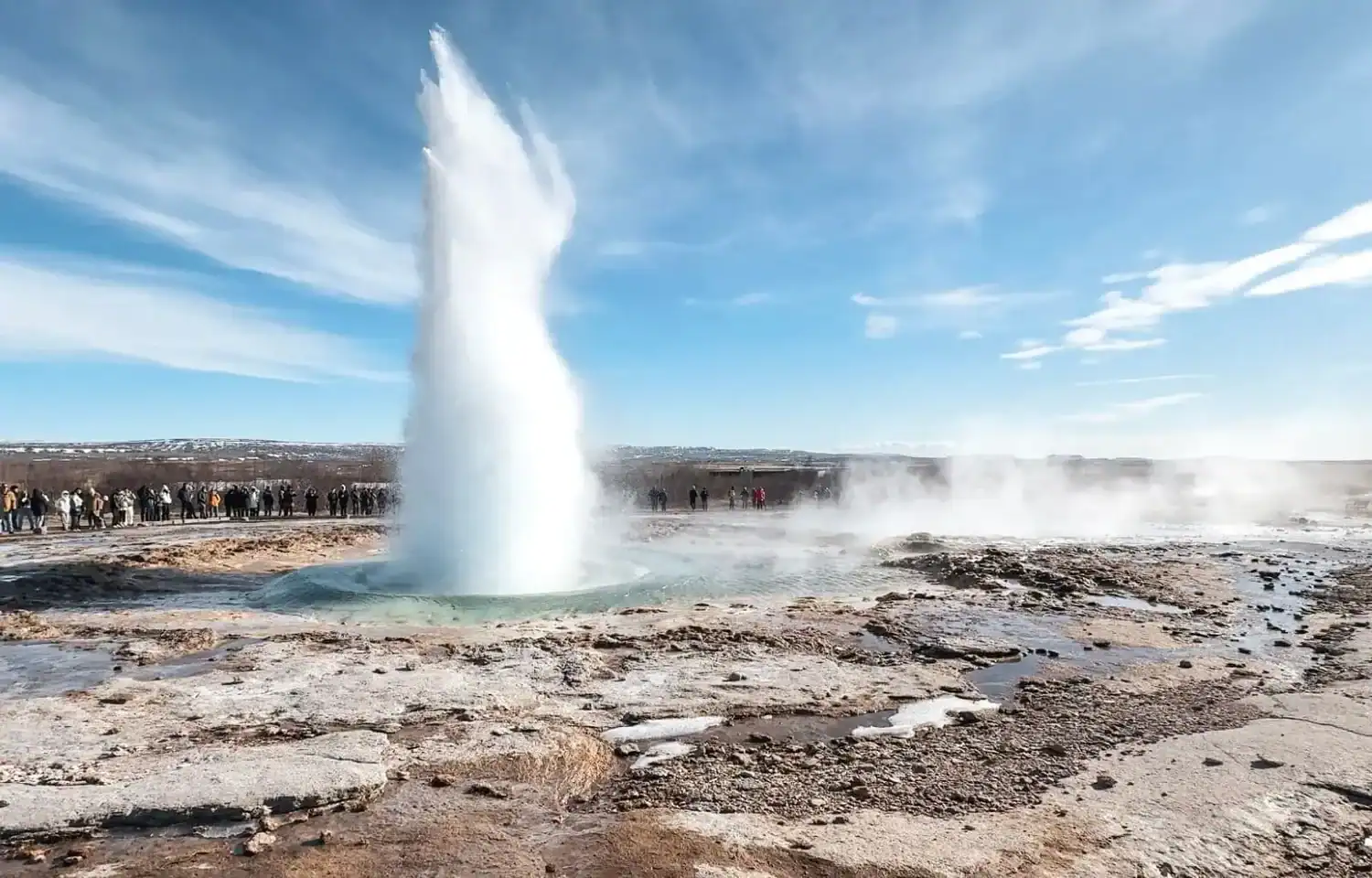 Powerful geyser erupting from a geothermal spring in Iceland, sending a tall column of water into the air beneath a bright blue sky. Tourists stand at a distance around the rocky landscape as steam rises across the surrounding geothermal area.