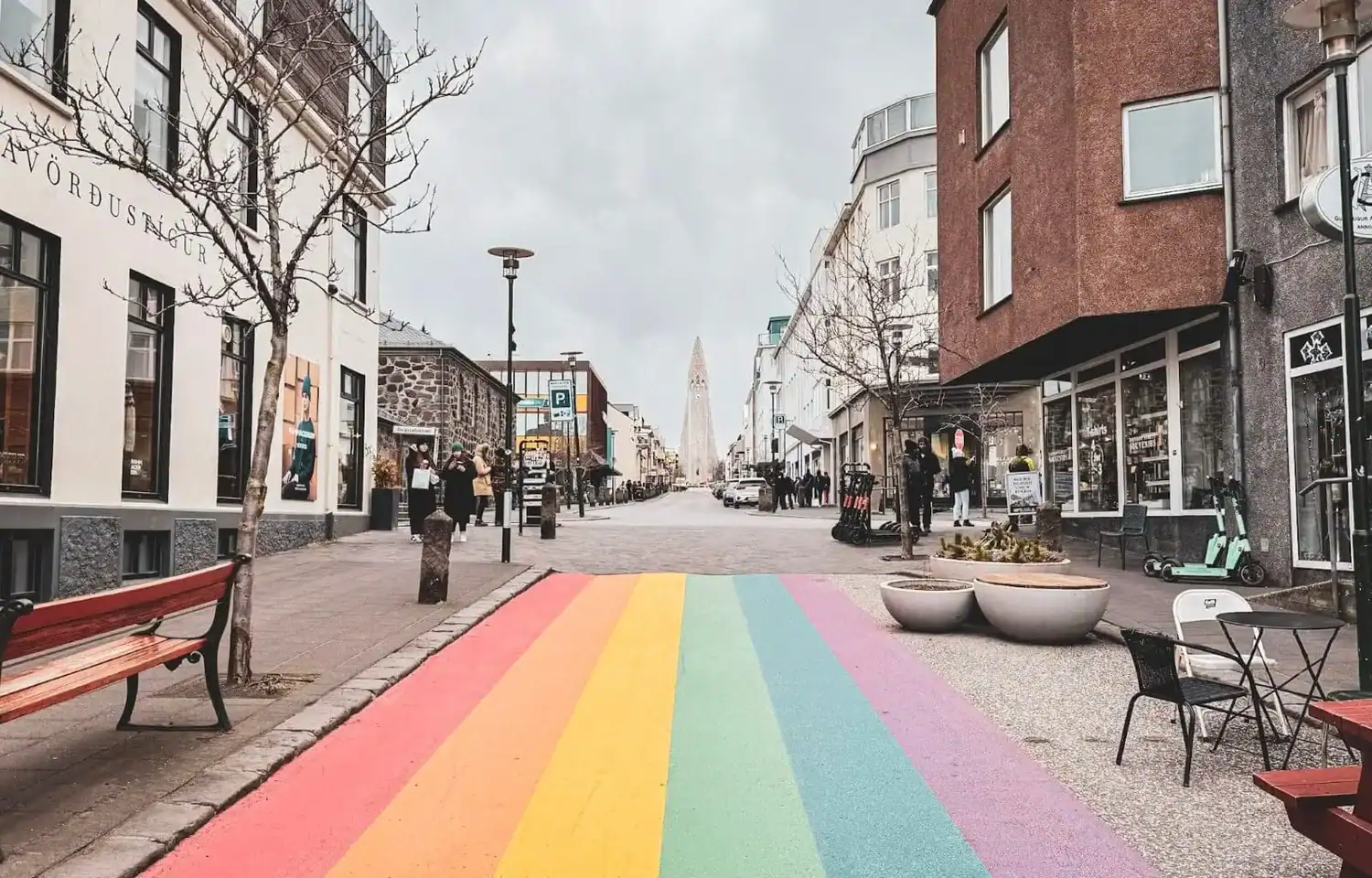 Rainbow-painted pedestrian crossing on a quiet city street lined with cafés and shops under an overcast sky. Bare trees, outdoor seating, and people walking along the pavement lead towards a tall church tower in the distance.
