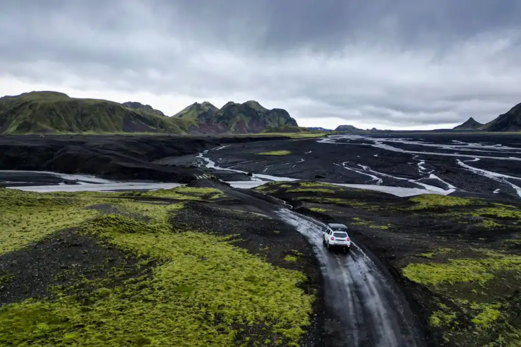 White 4x4 vehicle driving along a narrow dirt road through Iceland’s rugged highlands, surrounded by black volcanic plains, winding glacial rivers, and patches of bright green moss. Dark storm clouds hang low over distant mountains, creating a dramatic and remote landscape.