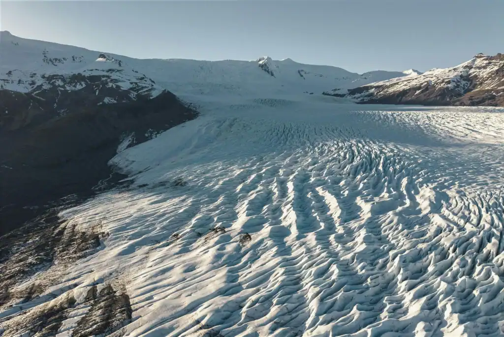 Snow-covered glacier flowing through a rugged mountain valley under a clear blue sky. Deep ridges and crevasses texture the icy surface, with dark volcanic rock and distant peaks framing the expansive frozen landscape.