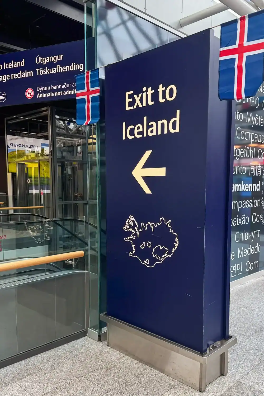 Large blue airport sign reading “Exit to Iceland” with a white arrow pointing left and an outline map of Iceland beneath. Icelandic flags hang above the sign inside a modern airport terminal with glass walls, escalators, and multilingual signage in the background.
