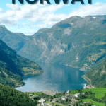 Scenic view of a fjord with steep green mountains and a small village at the water's edge under a partly cloudy sky.